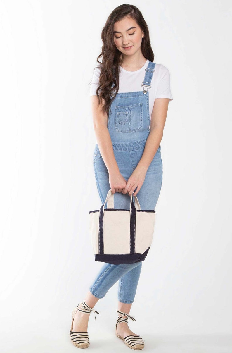 Puede incluir: Una mujer lleva un vestido de peto vaquero azul claro con una camiseta blanca y sandalias de esparto a rayas blancas y negras. Lleva un bolso de mano de lona blanca con ribetes negros.