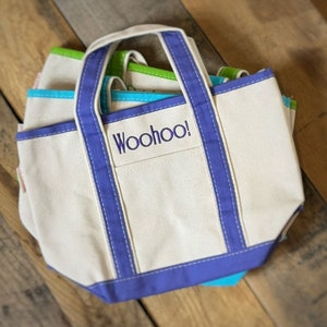 Pu&ograve; includere: Una pila di borse tote in tela di vari colori, tra cui bianco, blu e verde. La borsa anteriore ha un bordo viola e la parola "Woohoo!" ricamata sulla tasca anteriore. Le borse hanno manici robusti e un design classico.