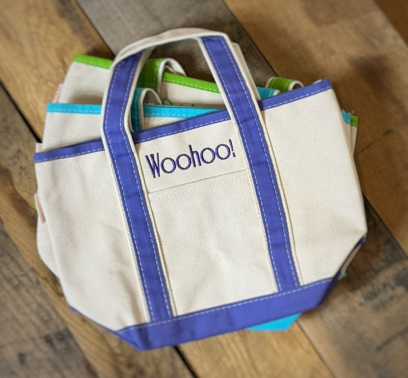 Pu&ograve; includere: Una pila di borse tote in tela di vari colori, tra cui bianco, blu e verde. La borsa anteriore ha un bordo viola e la parola "Woohoo!" ricamata sulla tasca anteriore. Le borse hanno manici robusti e un design classico.