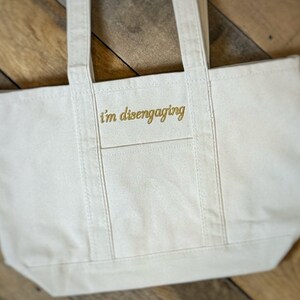 May include: A white canvas tote bag with the text "i'm disengaging" embroidered in gold on the front.