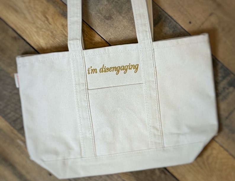 May include: A white canvas tote bag with the text "i'm disengaging" embroidered in gold on the front.
