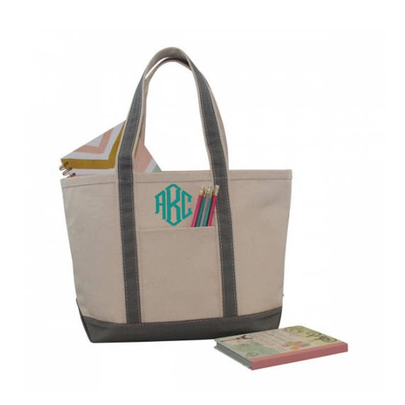 May include: A large, beige canvas tote bag with gray trim and a large pocket on the front. The bag has a monogram of the letters "ABC" in turquoise on the front pocket. The bag is filled with pencils and a notebook.
