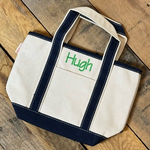 Puede incluir: Una peque&ntilde;a bolsa de lona blanca con ribetes y asas de color azul marino. La bolsa tiene un nombre bordado en verde "Hugh" en la parte delantera.