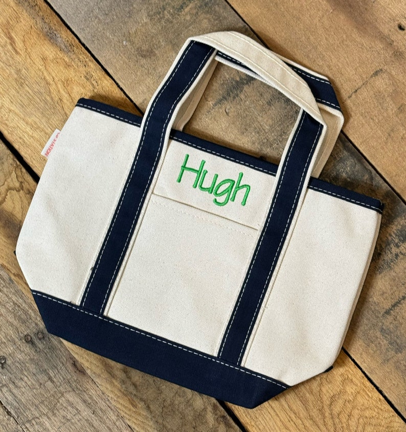 Puede incluir: Una peque&ntilde;a bolsa de lona blanca con ribetes y asas de color azul marino. La bolsa tiene un nombre bordado en verde "Hugh" en la parte delantera.