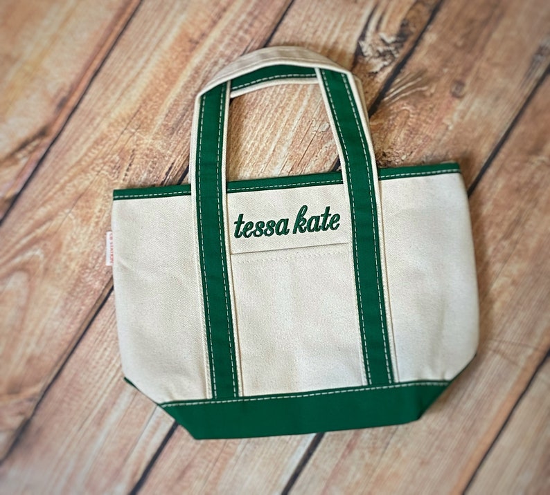 Puede incluir: Una peque&ntilde;a bolsa de lona blanca con ribetes verdes y el texto "tessa kate" bordado en verde en la parte delantera.