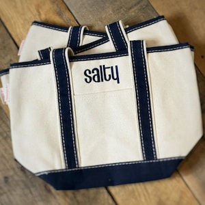 Pu&ograve; includere: Borse tote in tela bianco sporco con finiture e manici blu navy. La parola "salty" &egrave; ricamata in blu navy sulla tasca anteriore. Le borse sono impilate su una superficie di legno.