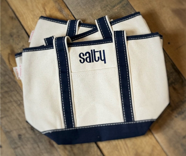 Pu&ograve; includere: Borse tote in tela bianco sporco con finiture e manici blu navy. La parola "salty" &egrave; ricamata in blu navy sulla tasca anteriore. Le borse sono impilate su una superficie di legno.