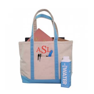 May include: A beige canvas tote bag with blue trim and a large pocket on the front. The bag is monogrammed with the letters "ASL" in red. The bag is filled with books and a water bottle. The water bottle is blue and white and says "JUST WATER" on the label.