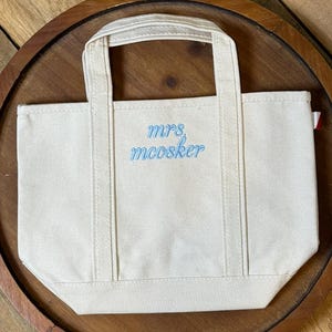 K&ouml;nnte beinhalten: Eine kleine cremewei&szlig;e Canvas-Tragetasche mit zwei Tragegriffen und einem flachen Boden. Auf der Vorderseite befindet sich der gestickte Text "mrs. mcosker" in hellblauer Schreibschrift. An der rechten Seitennaht ist ein kleines rot-wei&szlig;es Stoffetikett sichtbar.