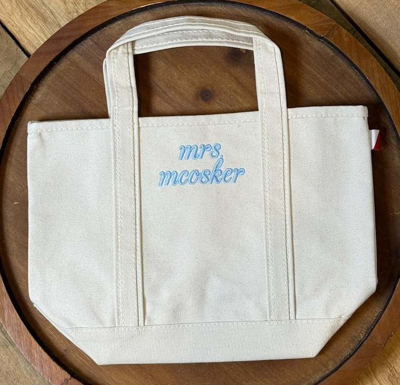 K&ouml;nnte beinhalten: Eine kleine cremewei&szlig;e Canvas-Tragetasche mit zwei Tragegriffen und einem flachen Boden. Auf der Vorderseite befindet sich der gestickte Text "mrs. mcosker" in hellblauer Schreibschrift. An der rechten Seitennaht ist ein kleines rot-wei&szlig;es Stoffetikett sichtbar.