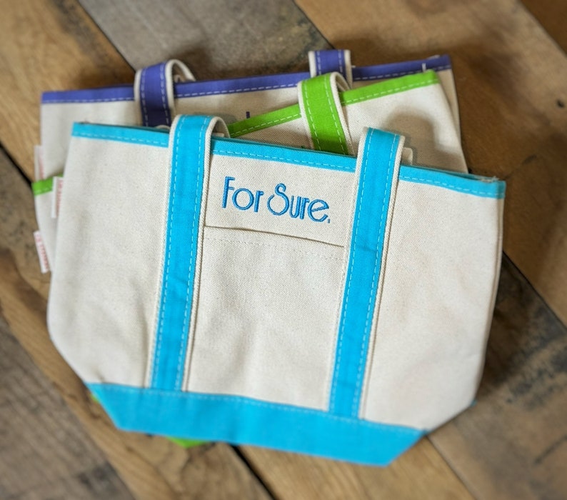 Pu&ograve; includere: Tre borse tote in tela con finiture e manici a contrasto blu, verdi e viola. Le borse sono bianco sporco con una piccola tasca frontale e la scritta ricamata "For Sure". Le borse sono su una superficie di legno.