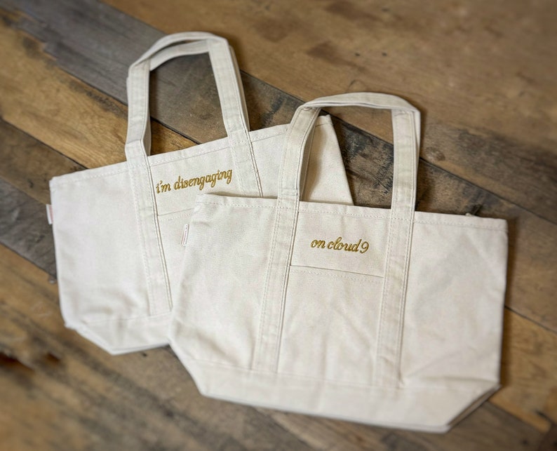Op de afbeelding: Twee witte canvas tassen met gouden geborduurde tekst. De tas aan de linkerkant zegt "i'm disengaging" en de tas aan de rechterkant zegt "on cloud 9".