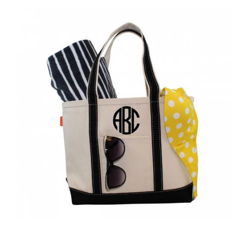 May include: A beige canvas tote bag with black trim and handles. The bag has a large black circle with the letters "ABC" in white. The bag is filled with a striped beach towel and a yellow polka dot swimsuit.