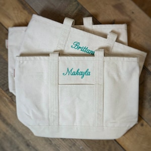 Op de afbeelding: Drie witte canvas tassen met turquoise geborduurde namen. De bovenste tas zegt "Brittani", de middelste tas zegt "Makayla", en de onderste tas is leeg.