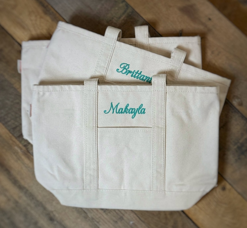 Op de afbeelding: Drie witte canvas tassen met turquoise geborduurde namen. De bovenste tas zegt "Brittani", de middelste tas zegt "Makayla", en de onderste tas is leeg.