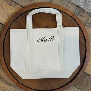K&ouml;nnte beinhalten: Eine kleine, cremewei&szlig;e Canvas-Tragetasche mit zwei stabilen Tragegriffen und einem verst&auml;rkten Boden. Auf der Vorderseite ist der Text "Mrs. R" in einer dunklen, eleganten Schreibschrift eingestickt, mittig zwischen den beiden vertikalen Tr&auml;gern.