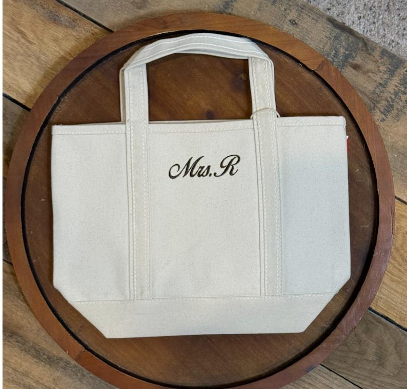 K&ouml;nnte beinhalten: Eine kleine, cremewei&szlig;e Canvas-Tragetasche mit zwei stabilen Tragegriffen und einem verst&auml;rkten Boden. Auf der Vorderseite ist der Text "Mrs. R" in einer dunklen, eleganten Schreibschrift eingestickt, mittig zwischen den beiden vertikalen Tr&auml;gern.