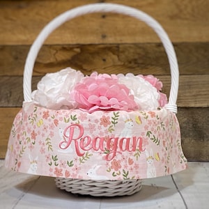 May include: White wicker basket with a pink and green floral fabric liner. The liner features white bunnies and butterflies. The basket is filled with pink and white tissue paper flowers. The fabric liner is personalized with the name "Reagan".
