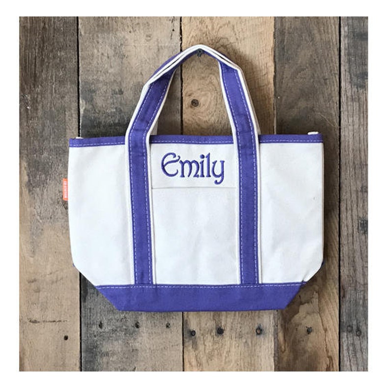 以下が含まれることがあります： 紫色の縁取りと正面に「Emily」と刺繍された白いキャンバス地のトートバッグです。