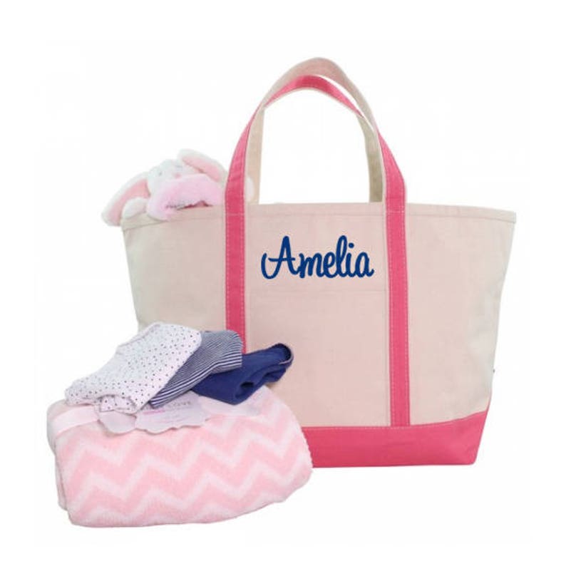 May include: A beige canvas tote bag with pink trim and the name "Amelia" embroidered in blue. The bag is filled with a pink and white chevron blanket and a stack of folded clothing.