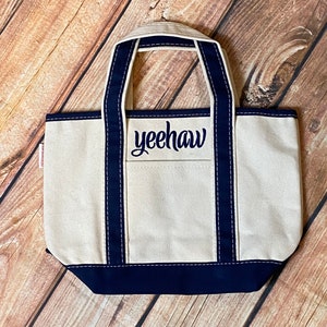May include: A white canvas tote bag with navy blue handles and trim. The bag has a front pocket with the word "yeehaw" embroidered in navy blue.
