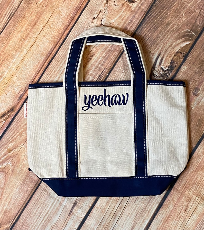 May include: A white canvas tote bag with navy blue handles and trim. The bag has a front pocket with the word "yeehaw" embroidered in navy blue.