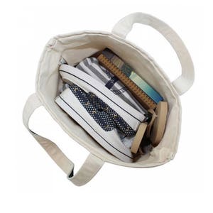 May include: A white canvas tote bag with two handles. The bag is filled with items, including a striped fabric, a book, and two pairs of shoes. The shoes are white with navy blue and gold accents.