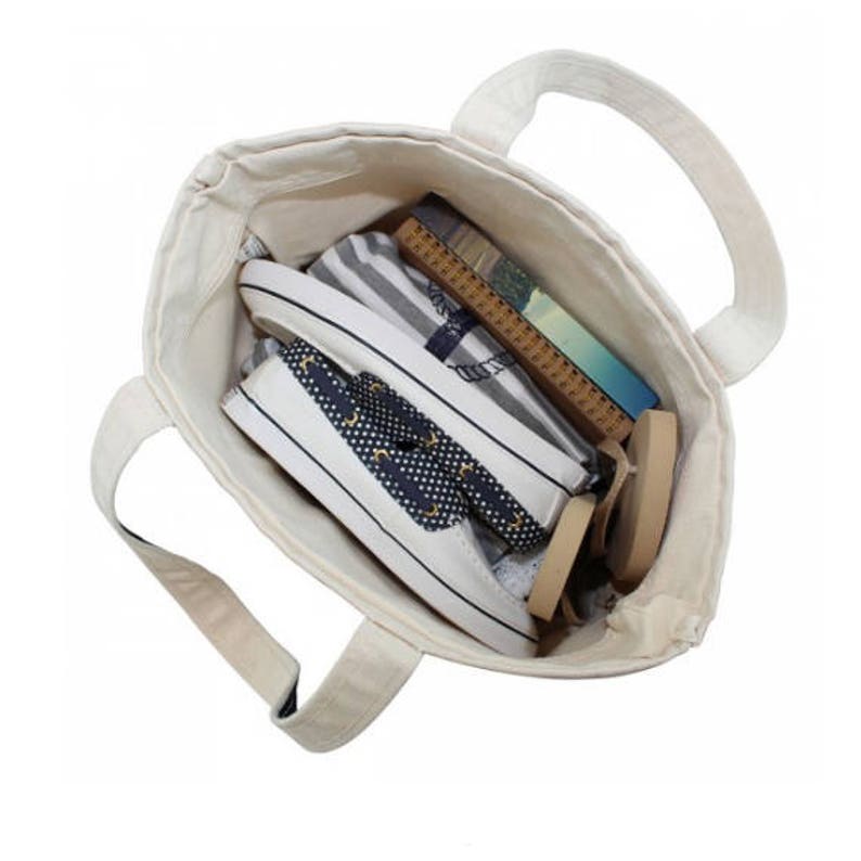 May include: A white canvas tote bag with two handles. The bag is filled with items, including a striped fabric, a book, and two pairs of shoes. The shoes are white with navy blue and gold accents.