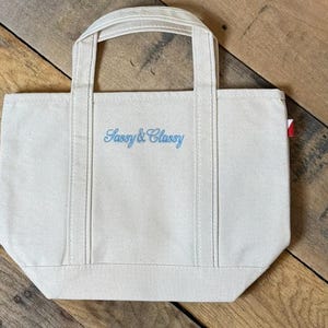 K&ouml;nnte beinhalten: Eine cremefarbene Canvas-Tragetasche mit zwei stabilen Griffen und einem flachen Boden. Auf der Vorderseite der Tasche befindet sich der gestickte Schriftzug "Sassy & Classy" in hellblauer Schreibschrift. Ein kleines rot-wei&szlig;es Etikett ist rechts sichtbar.