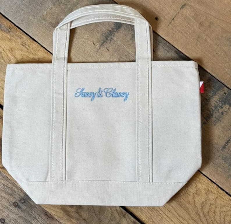 K&ouml;nnte beinhalten: Eine cremefarbene Canvas-Tragetasche mit zwei stabilen Griffen und einem flachen Boden. Auf der Vorderseite der Tasche befindet sich der gestickte Schriftzug "Sassy & Classy" in hellblauer Schreibschrift. Ein kleines rot-wei&szlig;es Etikett ist rechts sichtbar.