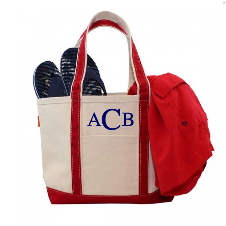 May include: A white canvas tote bag with red trim and blue embroidered initials "ACB". The bag is filled with a red shirt and blue flip flops.