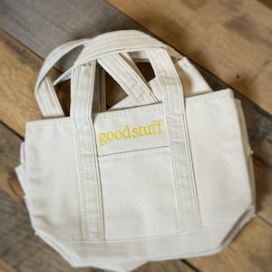 Pu&ograve; includere: Borsa tote in tela color crema con la scritta "good stuff" ricamata in giallo. La borsa ha una forma rettangolare, una piccola tasca e pi&ugrave; manici. &Egrave; appoggiata su una superficie di legno.