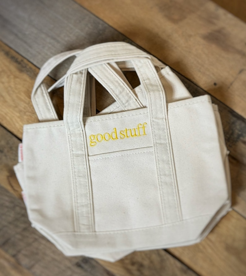 Pu&ograve; includere: Borsa tote in tela color crema con la scritta "good stuff" ricamata in giallo. La borsa ha una forma rettangolare, una piccola tasca e pi&ugrave; manici. &Egrave; appoggiata su una superficie di legno.