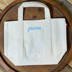 K&ouml;nnte beinhalten: Eine kleine, cremewei&szlig;e Canvas-Tragetasche mit zwei stabilen Tragegriffen und verst&auml;rkten vertikalen N&auml;hten. Der Name gianna ist in hellblauer Schreibschrift mittig auf der Vorderseite der Tasche eingestickt.