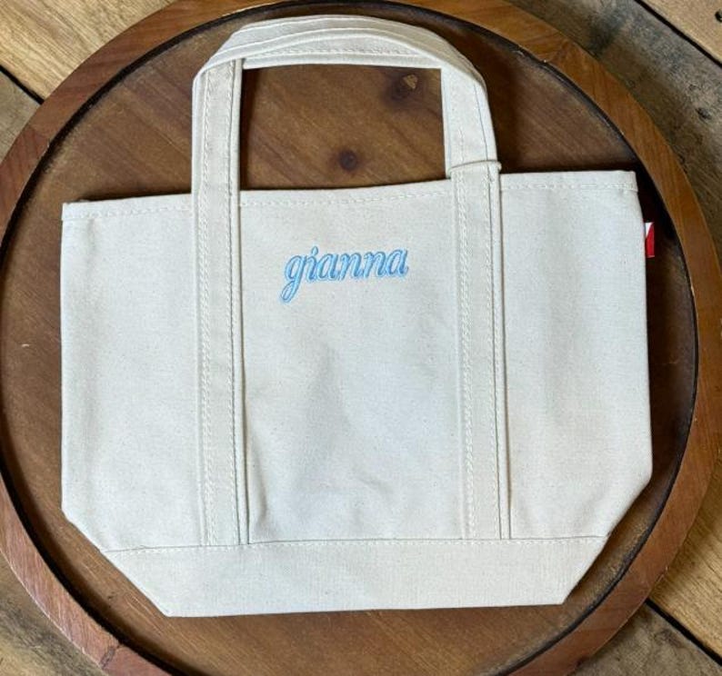 K&ouml;nnte beinhalten: Eine kleine, cremewei&szlig;e Canvas-Tragetasche mit zwei stabilen Tragegriffen und verst&auml;rkten vertikalen N&auml;hten. Der Name gianna ist in hellblauer Schreibschrift mittig auf der Vorderseite der Tasche eingestickt.
