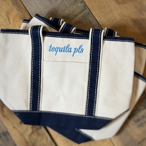 Pu&ograve; includere: Borse tote in tela bianco sporco con finiture e manici blu navy. Le borse hanno la scritta ricamata "tequila pls" in azzurro. Le borse sono impilate l'una sull'altra.