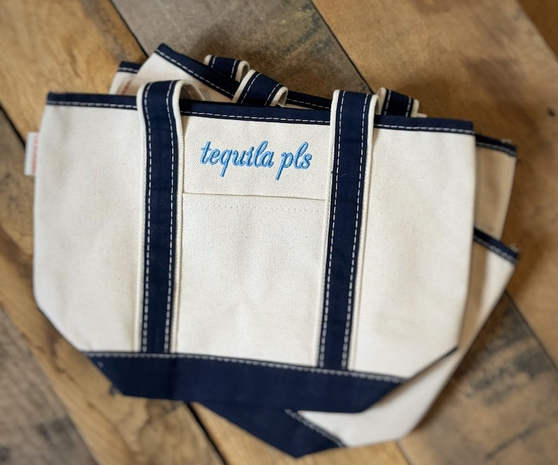 Pu&ograve; includere: Borse tote in tela bianco sporco con finiture e manici blu navy. Le borse hanno la scritta ricamata "tequila pls" in azzurro. Le borse sono impilate l'una sull'altra.