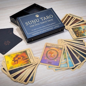 Può includere: Una scatola del mazzo di tarocchi Sunu Taro The Dog con un mazzo di carte. La scatola è blu navy con scritte dorate e un'impronta di zampa. Le carte illustrate sono a ventaglio su una superficie di legno.