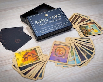 Puede incluir: Una caja de Sunu Taro The Dog Tarot Deck con un mazo de cartas. La caja es azul marino con letras doradas y una huella de pata. Las cartas ilustradas están dispuestas en abanico sobre una superficie de madera.