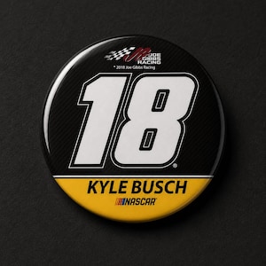 Peut inclure: Un bouton rond avec un fond noir en fibre de carbone affichant le numéro 18 en blanc. Le bouton a une bande jaune et noire en bas avec le texte "KYLE BUSCH" et "NASCAR". Le haut présente le logo Joe Gibbs Racing.