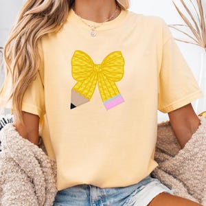 Könnte beinhalten: Hellgelbes T-Shirt mit gelber Schleife und Bleistift-Design. Der Bleistift hat eine schwarze Spitze und einen rosa Radiergummi. Das Shirt wird mit Jeansshorts und einem beigen Pullover getragen. Der Text unten im Bild lautet: Haftungsausschluss: Dies ist kein physisches Produkt, dies sind Vorschläge zur Verwendung dieses Bundles.