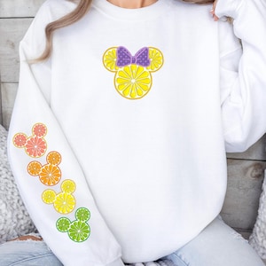 Peut inclure: Sweat-shirt blanc avec un motif sur le thème des agrumes. Le devant présente un motif inspiré de Mickey Mouse fait de tranches de citron avec un nœud violet. La manche gauche affiche des motifs d'agrumes en orange, pamplemousse, citron et citron vert. Le texte de l'avertissement est en bas.