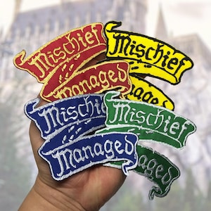 Può includere: Toppe ricamate rosse, gialle, blu e verdi, ognuna con le parole "Mischief Managed" in un carattere stilizzato. Le toppe sono tenute in mano, con un castello sfocato sullo sfondo.