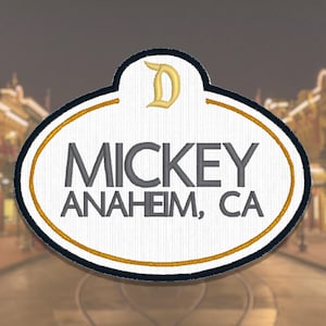 May include: An oval-shaped embroidered patch with a white background and a black border. The patch features a gold letter D at the top and the text MICKEY ANAHEIM, CA in charcoal gray lettering. A thin gold line circles the interior of the patch.
