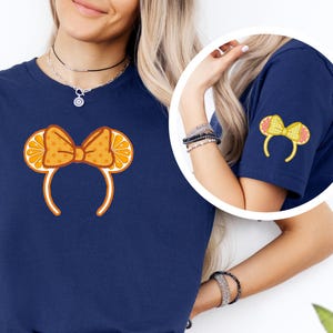 Peut inclure: T-shirt bleu marine avec un motif de serre-tête sur le thème des oranges. Le serre-tête présente un nœud avec des tranches d'orange et un motif à pois. Une version plus petite du motif est sur la manche. Le t-shirt est porté avec un jean bleu.