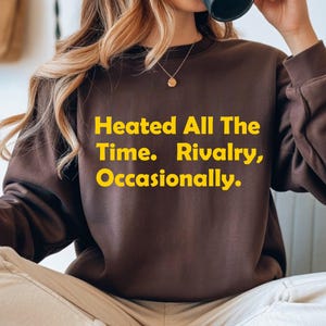 Può includere: Una felpa marrone con la scritta gialla "Heated All The Time. Rivalry, Occasionally.". La felpa ha maniche lunghe e girocollo. La persona tiene in mano una tazza nera.