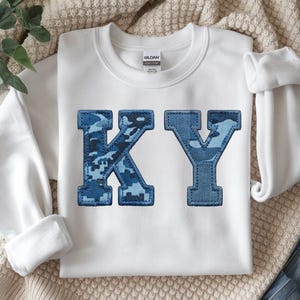 May include: White sweatshirt with the letters "KY" appliqued on the front. The letters are blue denim with a camouflage pattern. The sweatshirt has long sleeves and a crew neck.