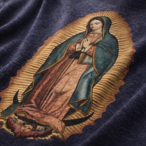 May include: Close-up of a navy blue fabric featuring a detailed print of the Virgin Mary. The image depicts the Virgin Mary with a halo, wearing a blue cloak and pink robe. The artwork has a vintage, religious theme.