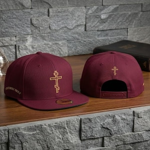 Op de afbeelding: Twee bordeauxrode baseballpetten met gouden kruisen. Eén pet wordt van voren bekeken en toont het kruis en de tekst "MOST HOLY HOLY". De andere pet wordt van achteren bekeken en toont ook een gouden kruis. De petten liggen op een houten oppervlak.