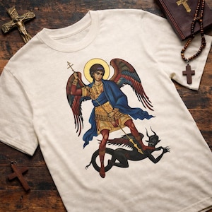 May include: Cream-coloured t-shirt with a detailed illustration of an angel with a halo, holding a cross and spear, battling a black demon. The angel has red, blue, and gold accents. The shirt is made of a soft material.